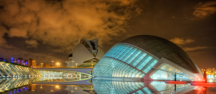 A photo of several highly stylized buildings lit at night in multiple colors, with their mirror reflection in water.