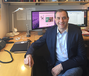 Image of a man sitting in a chair at a desk, looking at the camera. A computer is in the background.