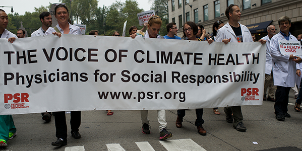 Physicians in white coats holding a banner that reads “The Voice of Climate Health: Physicians for Social Responsibility.” Others hold signs that read “Climate Change is a Health Risk.” 