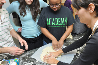 The teacher and several students stand around a counter watching the teacher spread out the pizza dough.