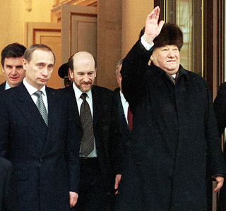 A grey-haired gentleman wearing a fur hat and black overcoat, raises his hand in a wave.