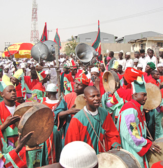 Photo of many people gathered in a street, with large loudspeakers, hand drums, and a hand holding up a tape recorder.