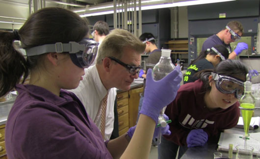 Two female students stand at a lab bench with a male professor. They are looking at a glass container filled with a neon yellow liquid.