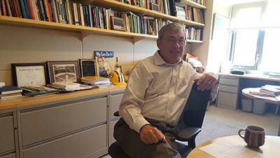 Smiling man sitting in a chair in an office. Filing cabinets and bookshelves are positioned behind the chair.