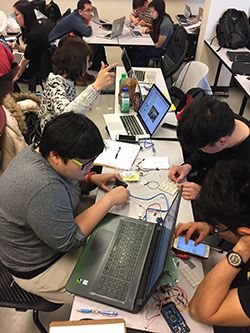 Several students sitting around a rectangular table. They’re manipulating electronics.